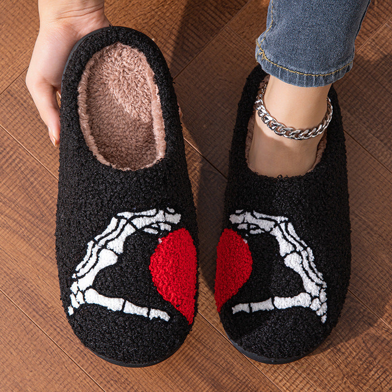 Black slippers with skeleton and heart design held by a person on a wooden floor.